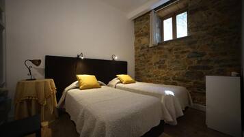 A bedroom with two beds, a stone wall, a window, and a table with a lamp.