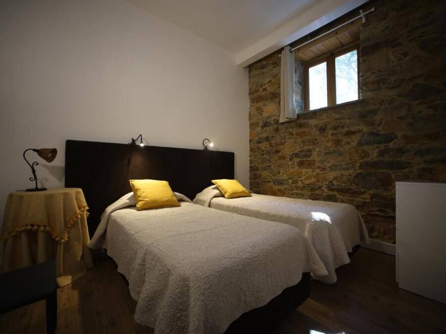 A bedroom with two beds, a stone wall, a window, and a table with a lamp.