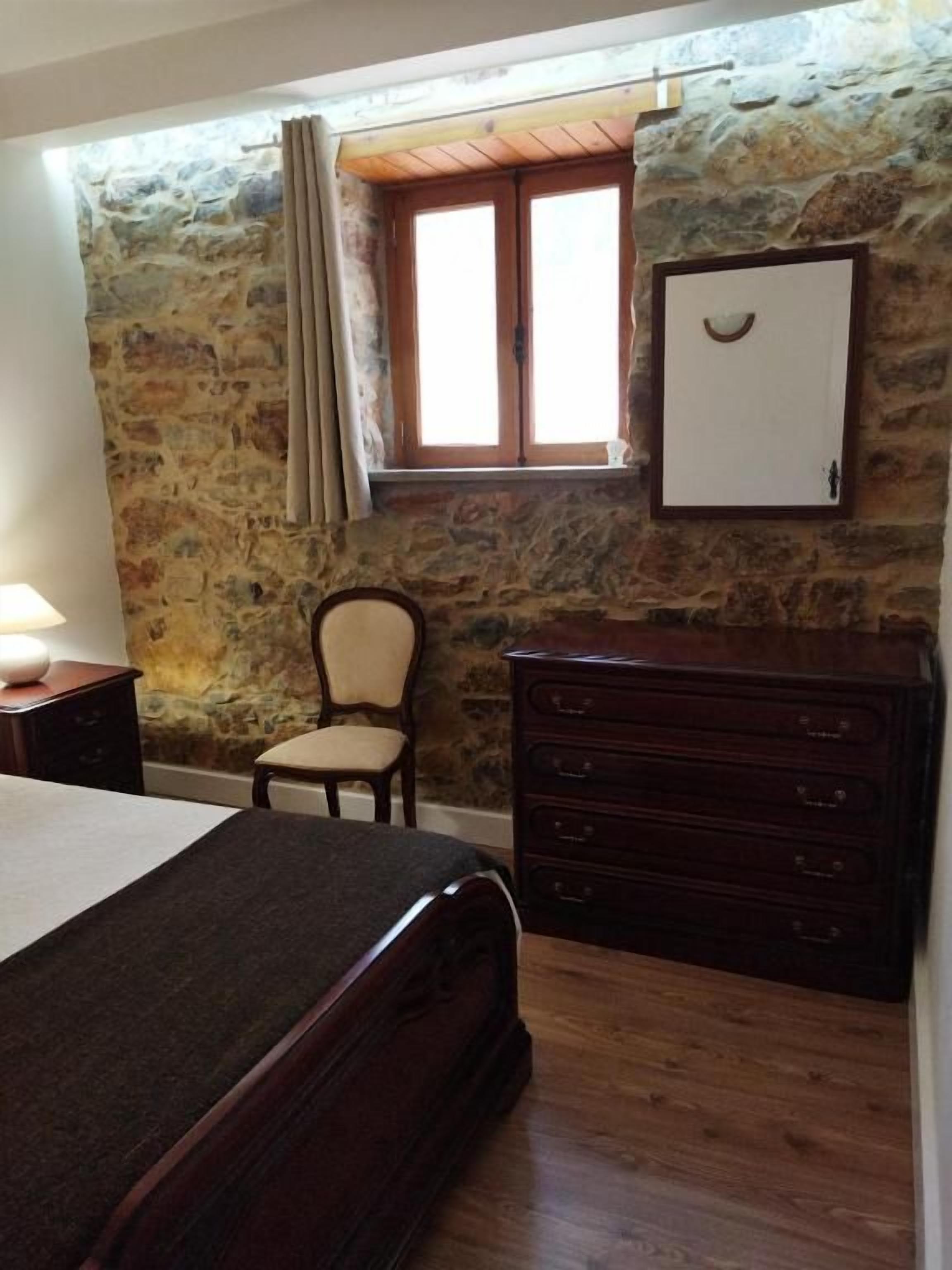 A bedroom with a stone wall, a wooden dresser, a bed, a chair, and a window with curtains.
