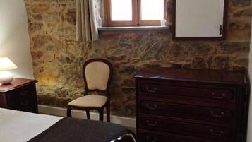 A bedroom with a stone wall, a wooden dresser, a bed, a chair, and a window with curtains.