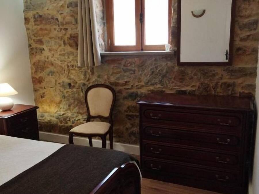 A bedroom with a stone wall, a wooden dresser, a bed, a chair, and a window with curtains.
