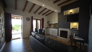 Sala de estar com sofá de couro preto, vigas de madeira no teto, área de cozinha com armários e micro-ondas, e uma lareira.
