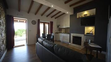 A living room with a black leather sofa, wooden beams on the ceiling, a kitchen area with cabinets and a microwave, and a fireplace.