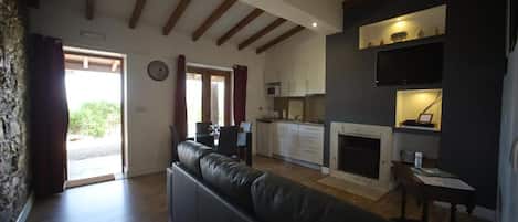 Una sala de estar con un sofá de cuero negro, vigas de madera en el techo, una zona de cocina con armarios y un microondas, y una chimenea.