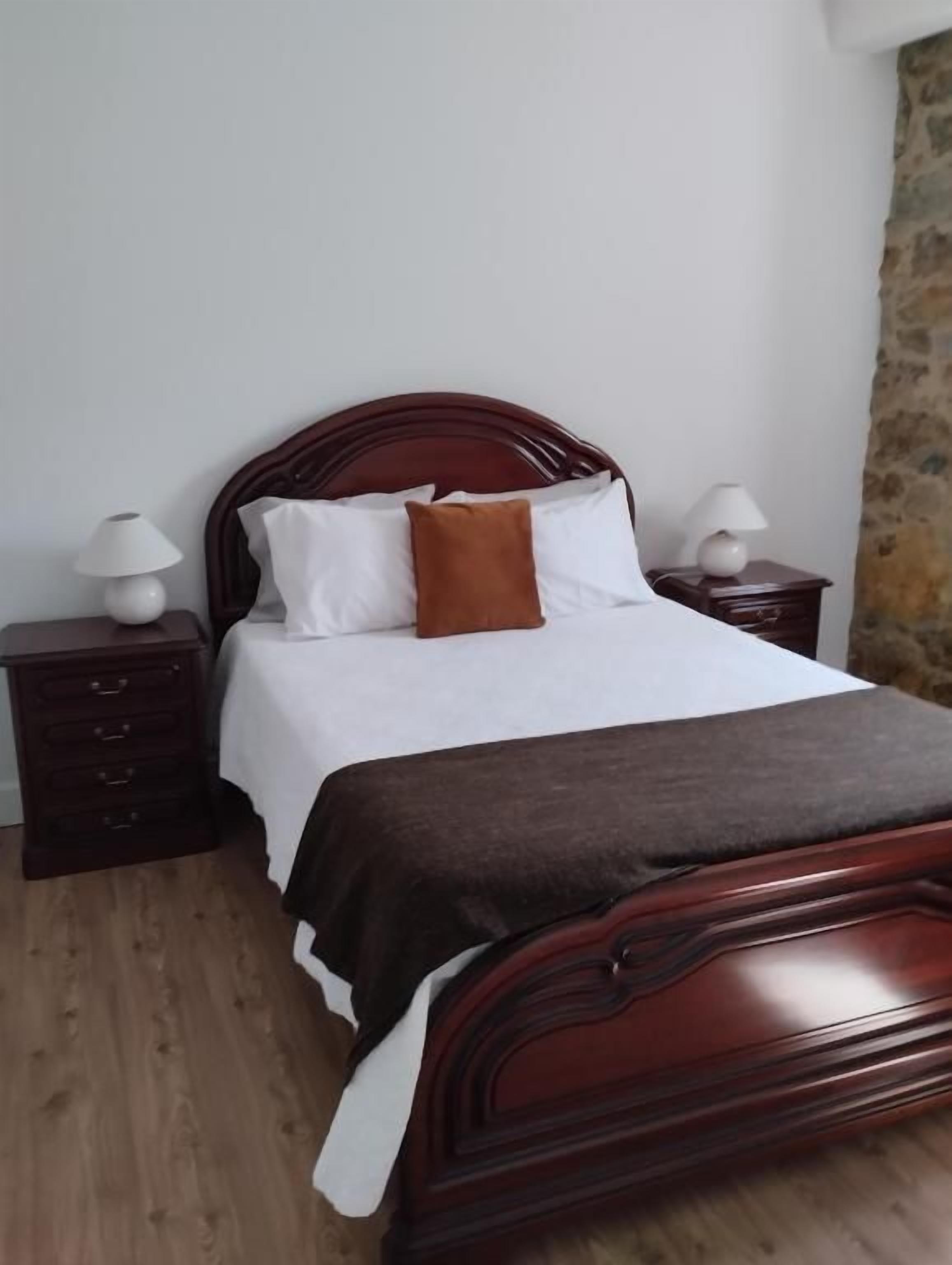 A single bed with a wooden headboard, white bedding, and a brown throw pillow. There are two bedside tables with lamps. The room has a stone wall and wooden flooring.