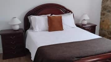A single bed with a wooden headboard, white bedding, and a brown throw pillow. There are two bedside tables with lamps. The room has a stone wall and wooden flooring.
