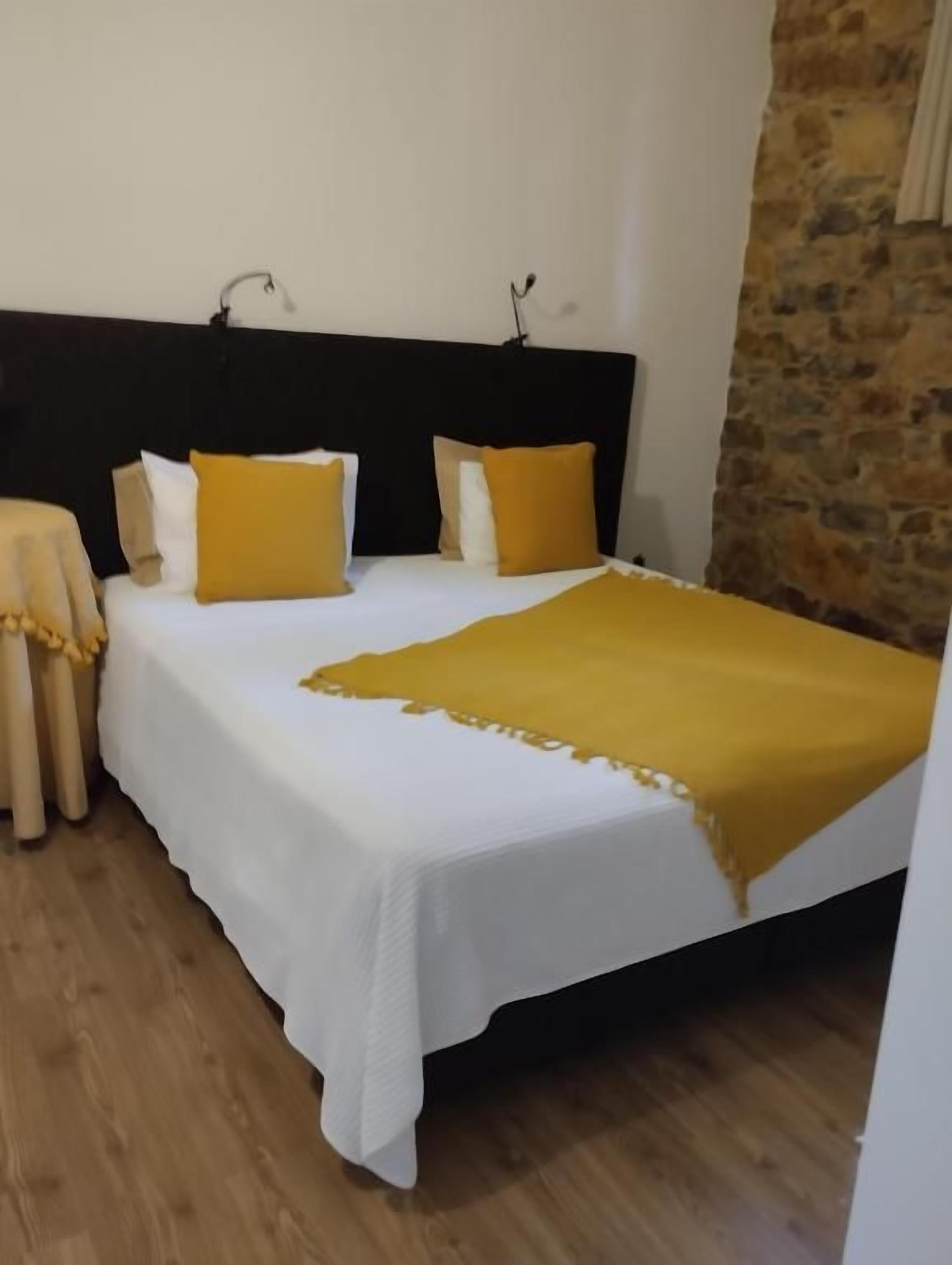 A bed with a white bedspread and a yellow throw, a dark headboard, and a stone wall.