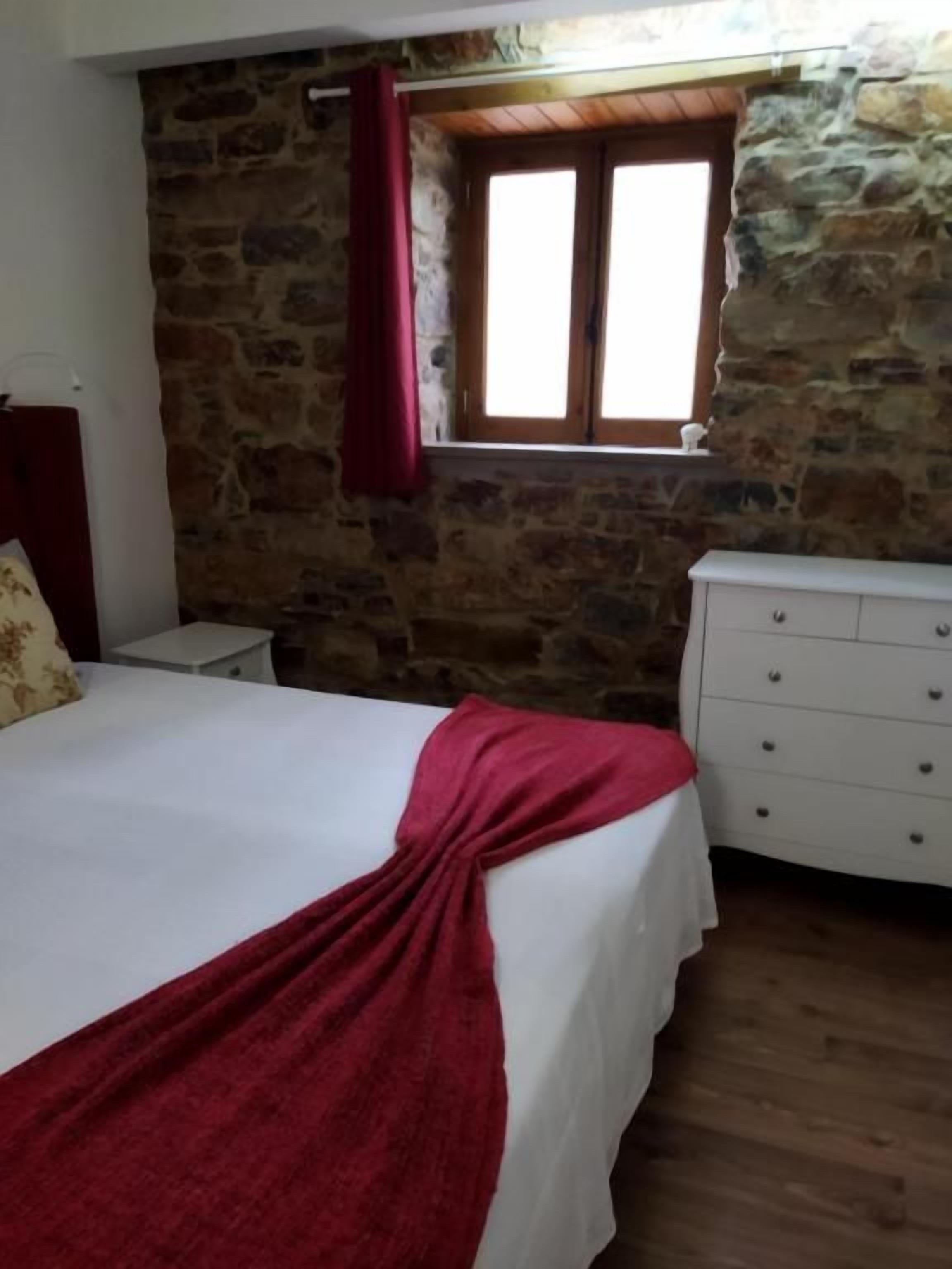 A bedroom with a stone wall, a window with wooden shutters, a bed with a white bedspread, and a white dresser.