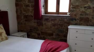 A bedroom with a stone wall, a window with wooden shutters, a bed with a white bedspread, and a white dresser.