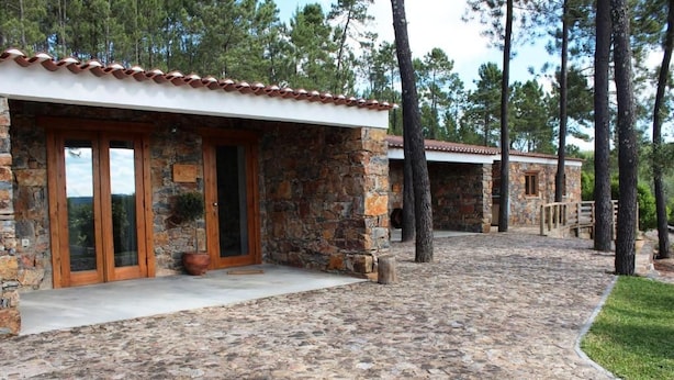 Um edifício de pedra com telhado em tijolos, entrada pavimentada e um caminho que leva a outra construção.