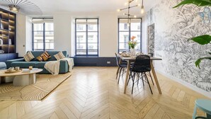 A modern living room with a sofa, coffee table, dining table, and chairs. There are large windows, a patterned wall, and a ceiling light fixture. - FurtiveStay - Sentier - 5 Jeuneurs - 2BDR 6P (Paris)