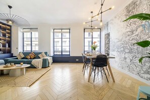 Ein modernes Wohnzimmer mit einer Couch, einem Couchtisch, einem Esstisch und Stühlen. Es gibt große Fenster, eine strukturierte Wand und eine Deckenleuchte.