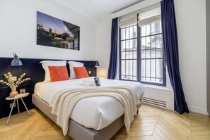 Ein Hotelzimmer mit Bett, Nachttisch, Lampe, einem Fenster mit Vorhängen und einem Stadtbild an der Wand.
