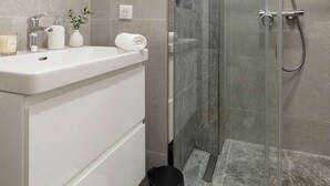A modern bathroom with a glass shower enclosure, a white vanity with a mirror, and a small trash bin. - FurtiveStay - Sentier - 5 Jeuneurs - 2BDR 6P (Paris)