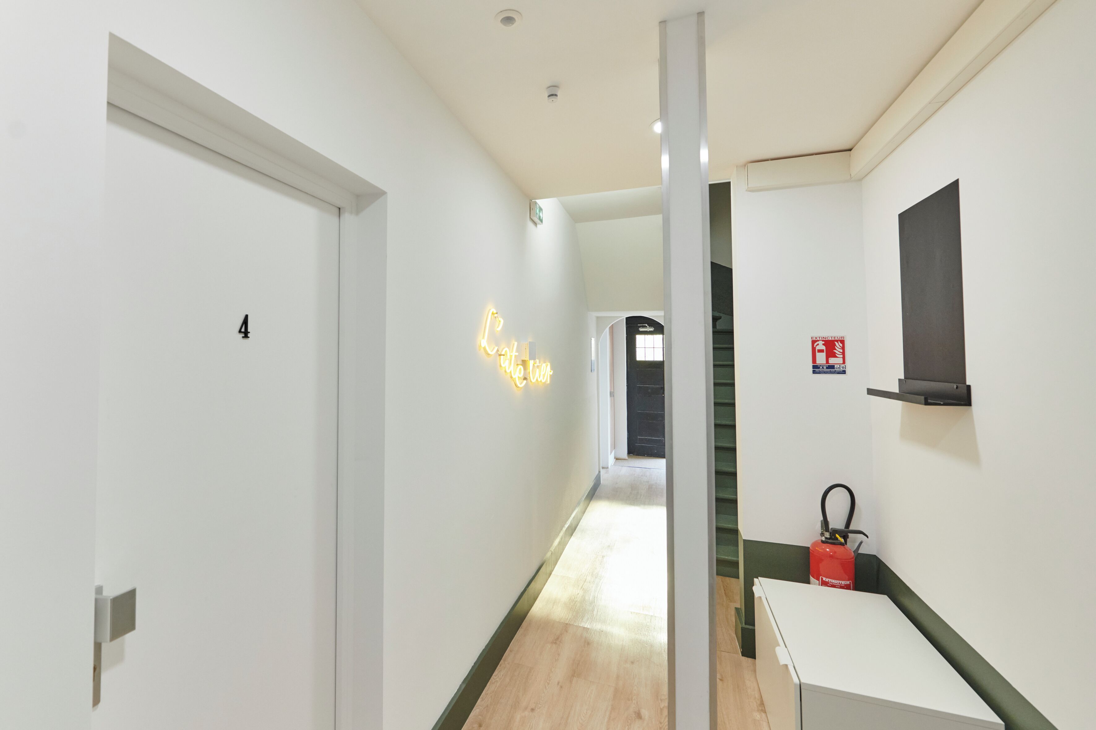 Un couloir avec une porte blanche portant le numéro « 4 », un panneau lumineux au néon, un extincteur et un escalier.
