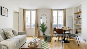 A living room with a sofa, coffee table, dining table with chairs, a potted plant, and a view through large windows. - FurtiveStay - Opéra - Hanovre - 1BDR 4P (Paris)