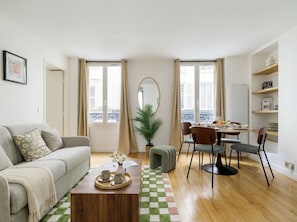 Ein Wohnzimmer mit Sofa, Couchtisch, Esstisch mit Stühlen, einer Topfpflanze und Blick durch große Fenster.