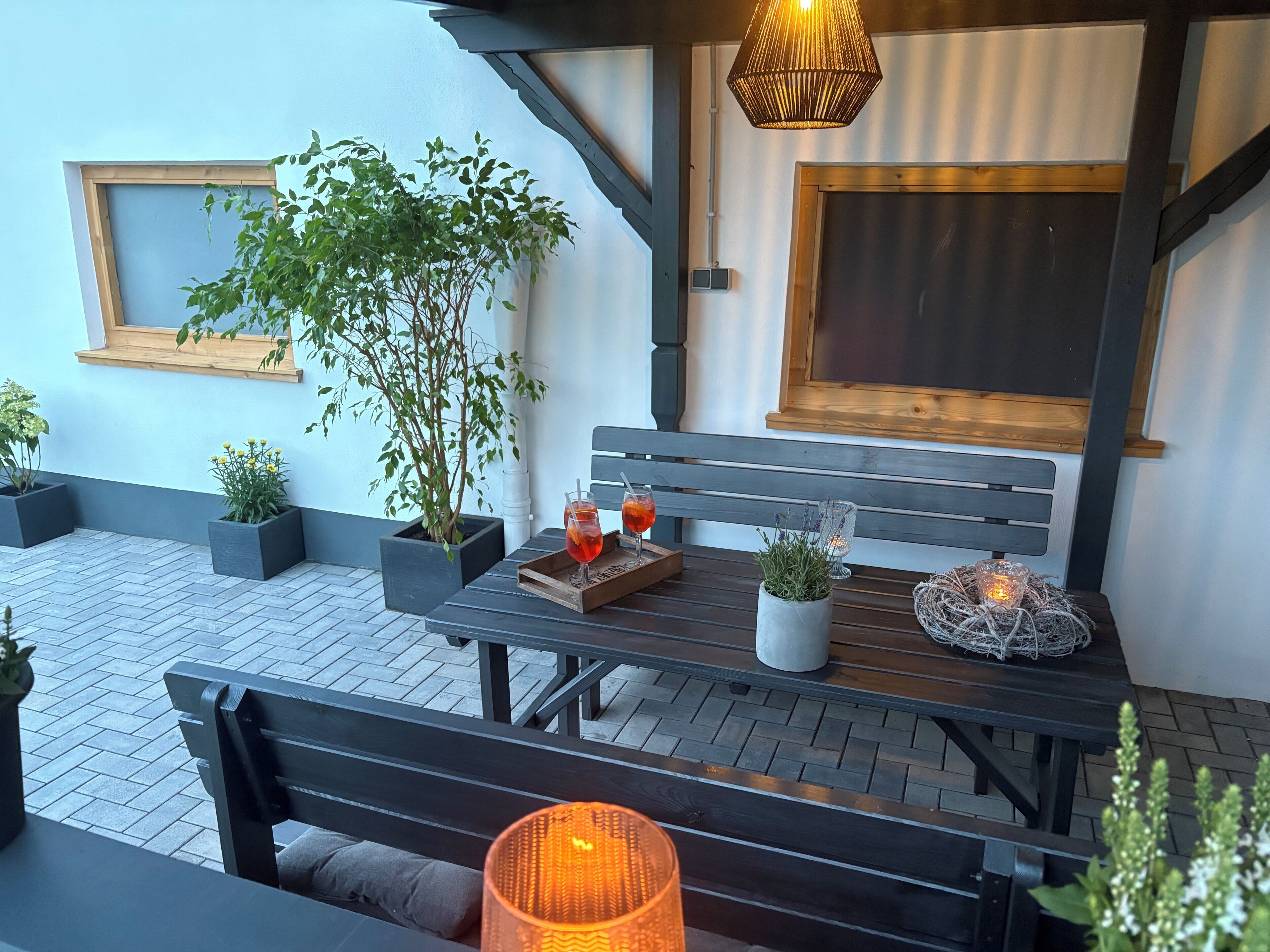 Eine Terrasse mit einer Holzbank, einem Tisch mit Getränken, Topfpflanzen und einer hängenden Leuchte.