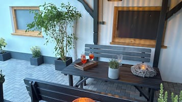 Eine Terrasse mit einer Holzbank, einem Tisch mit Getränken, Topfpflanzen und einer hängenden Leuchte.