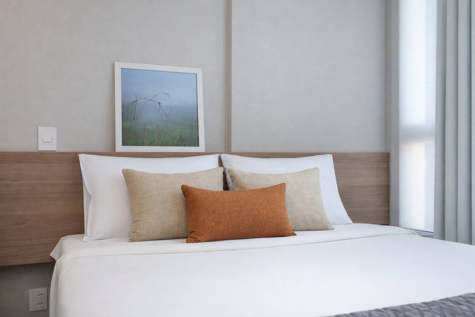 A neatly made bed with white bedding and two pillows, one brown and one beige, in a room with a plain wall and a window with curtains.