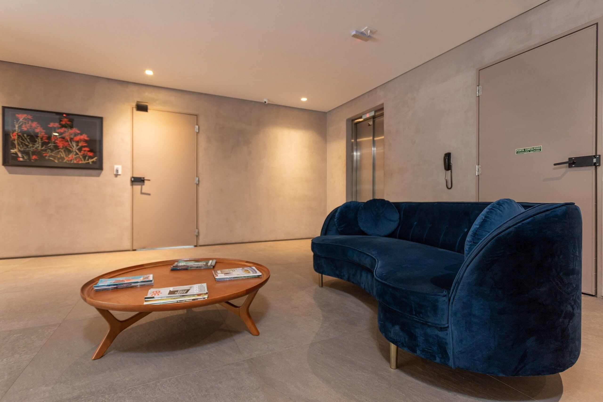A modern lobby with a blue velvet sofa, a wooden coffee table with magazines, a framed picture on the wall, and an elevator.