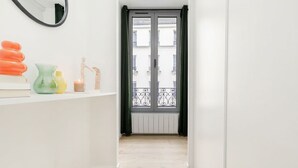 Ein Flur mit weißer Wand, einem Spiegel, einer kleinen Ablage mit Dekoration und einer Tür, die auf einen Balkon mit Metallgeländer führt.