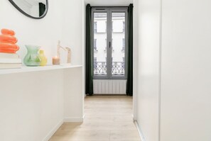 Ein Flur mit weißer Wand, einem Spiegel, einer kleinen Ablage mit Dekoration und einer Tür, die auf einen Balkon mit Metallgeländer führt.