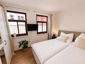 Ein Hotelzimmer mit einem Bett, einem an der Wand montierten Fernseher, einer Topfpflanze und Jalousien an den Fenstern.