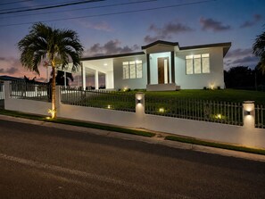 A modern two-story house with a covered porch, a fenced yard, and a well-lit entrance. - Casa Cecilia Retreat w/pool Near PR’s Best Beaches (Aguada)