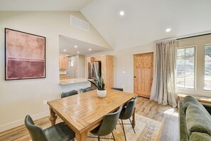 A dining area with a wooden table and chairs, a kitchen in the background, and a large window with curtains. - Brand New Cozy Getaway - Mountain Views + Firepit (Buena Vista)