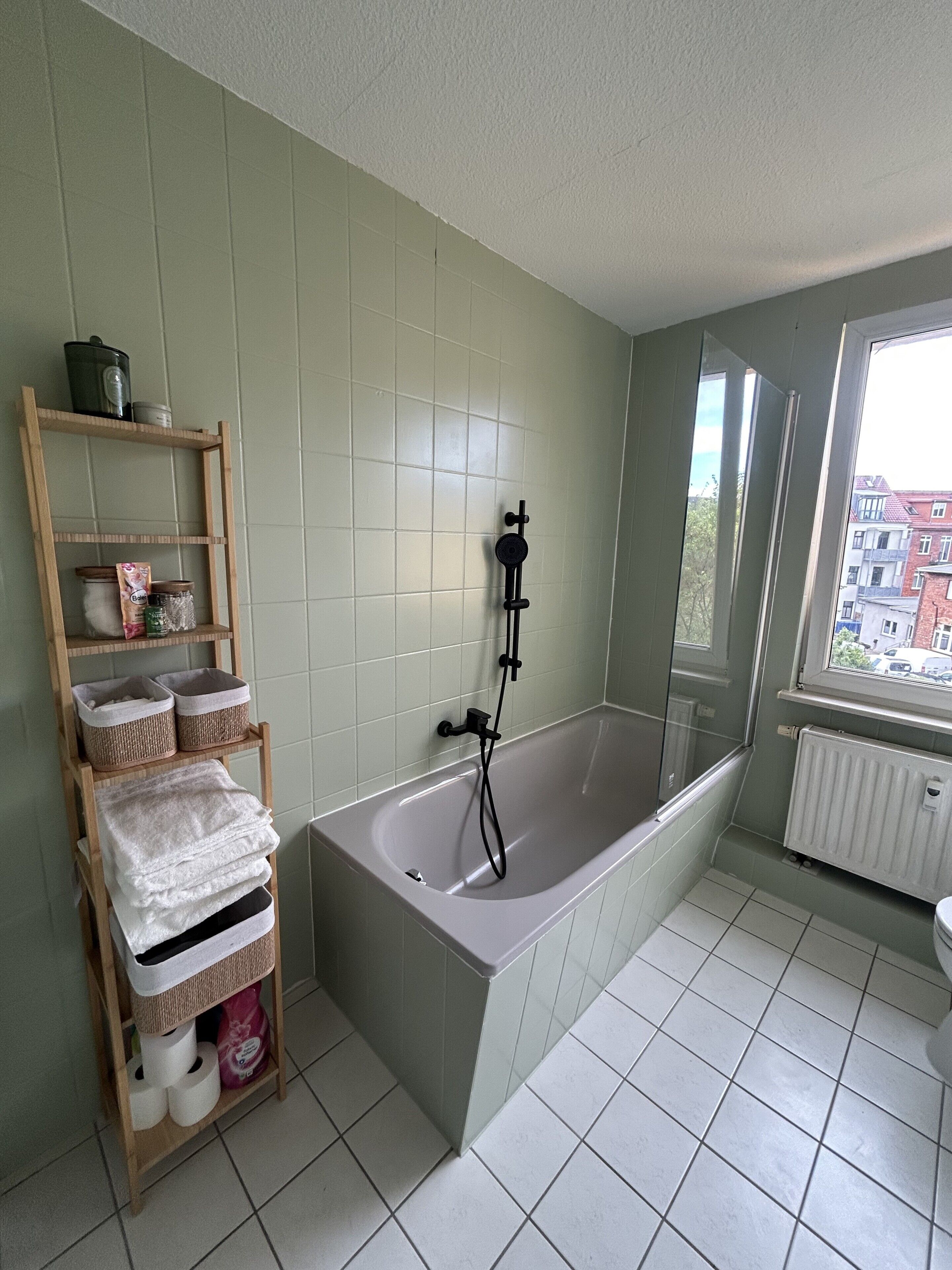 Ein Badezimmer mit einer Badewanne, einem hölzernen Regal mit Körben und einem Fenster mit Blick auf Gebäude.