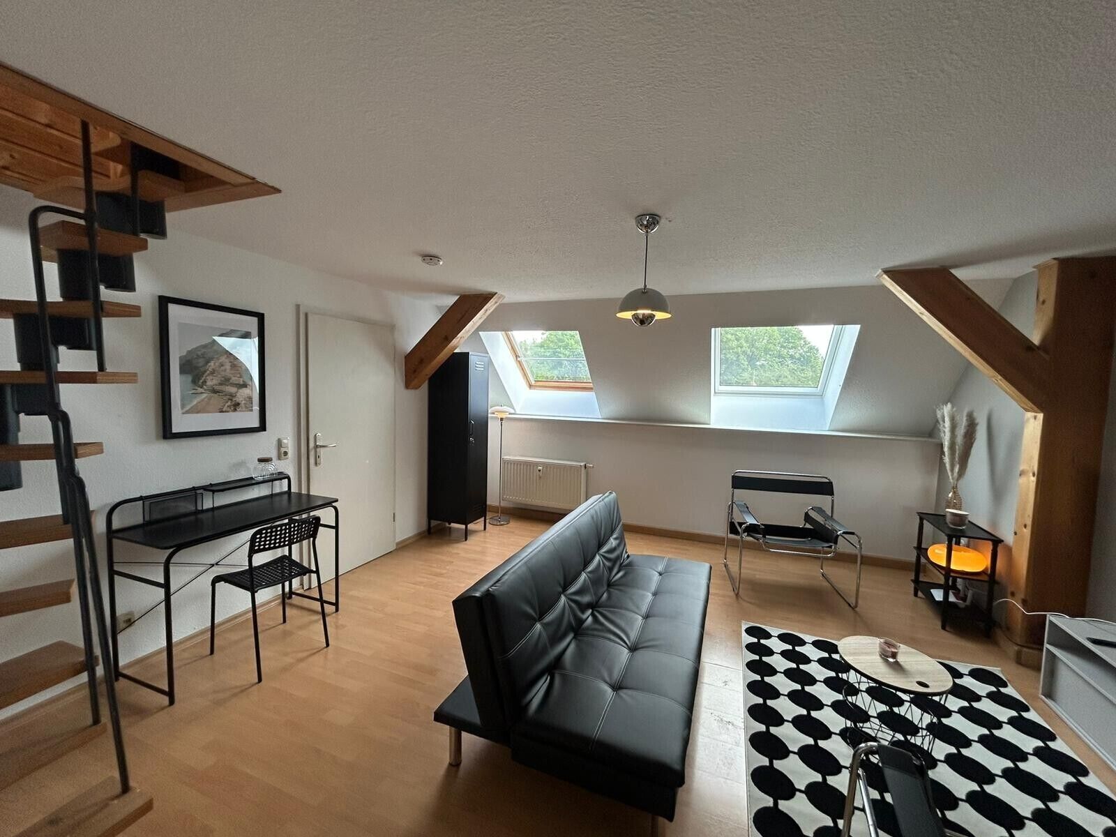 Een loft-achtige kamer met een zwarte leren bank, een eettafel met stoelen, een zwart metalen bureau, een radiator en dakramen.