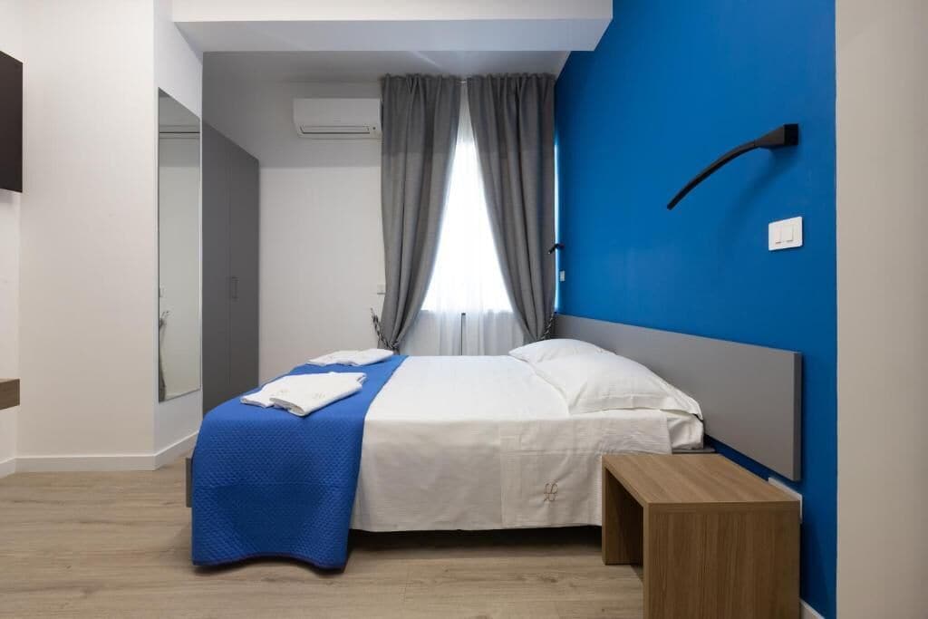 A bedroom with a bed, a bedside table, a window with curtains, and a wall-mounted light fixture.