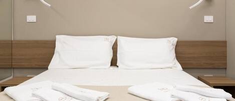A neatly made bed with white linens and towels, a wooden headboard, and a wall-mounted light fixture.