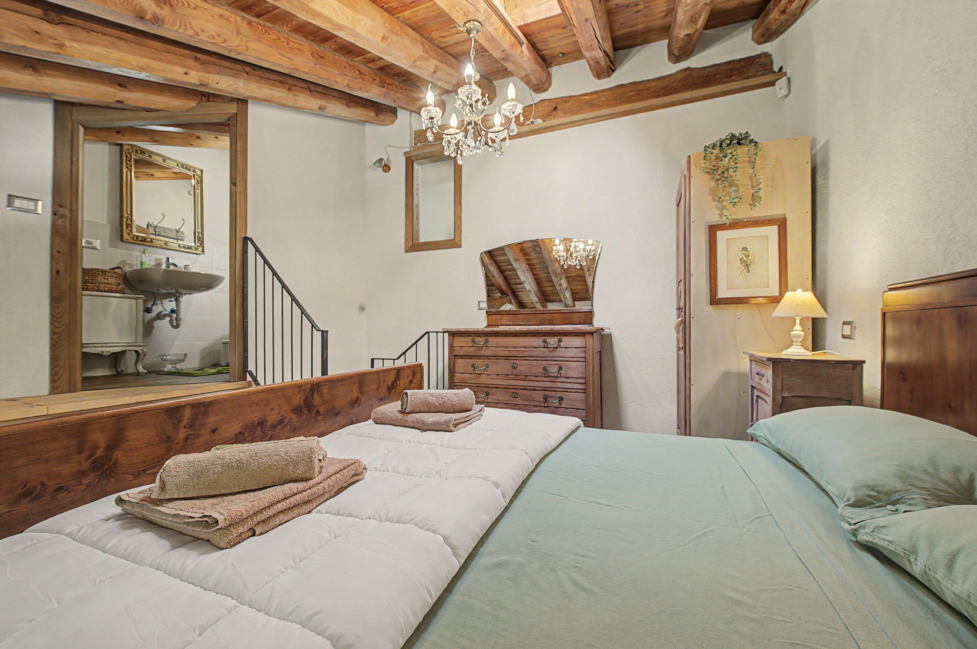 Une chambre avec un plafond en bois, un lit avec une literie blanche, une commode en bois, un miroir et un lustre.
