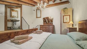 Une chambre avec un plafond en bois, un lit avec une literie blanche, une commode en bois, un miroir et un lustre.
