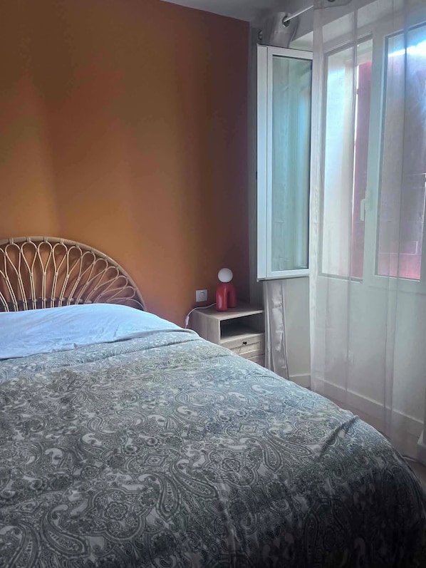 A bedroom with a bed, a nightstand, a window with curtains, and a view of the outside.