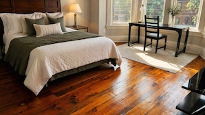 Quarto com piso de madeira, cama com cabeceira, mesa de cabeceira com abajur, janela com vista para prédios e cadeira perto da janela.