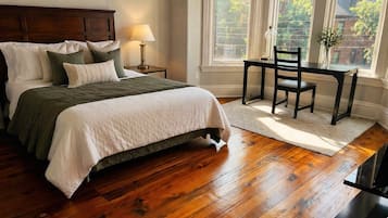 A bedroom with a wooden floor, a bed with a headboard, a nightstand with a lamp, a window with a view of buildings, and a chair near the window.