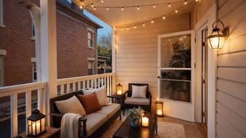 A covered porch with a sofa, chairs, and tables, string lights, and a door with a view of greenery.