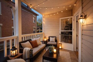 Une véranda aménagée avec un canapé, des fauteuils et des tables, des guirlandes lumineuses et une porte donnant sur un paysage verdoyant.