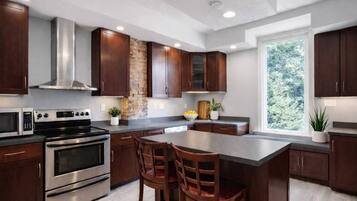 A modern kitchen with dark wood cabinets, stainless steel appliances, a central island with seating, and a large window providing a view of greenery.
