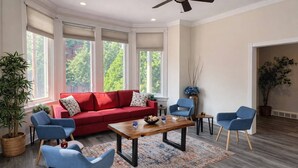 Uma sala de estar moderna com um sofá vermelho, cadeiras azuis, uma mesa de centro de madeira e grandes janelas com persianas.