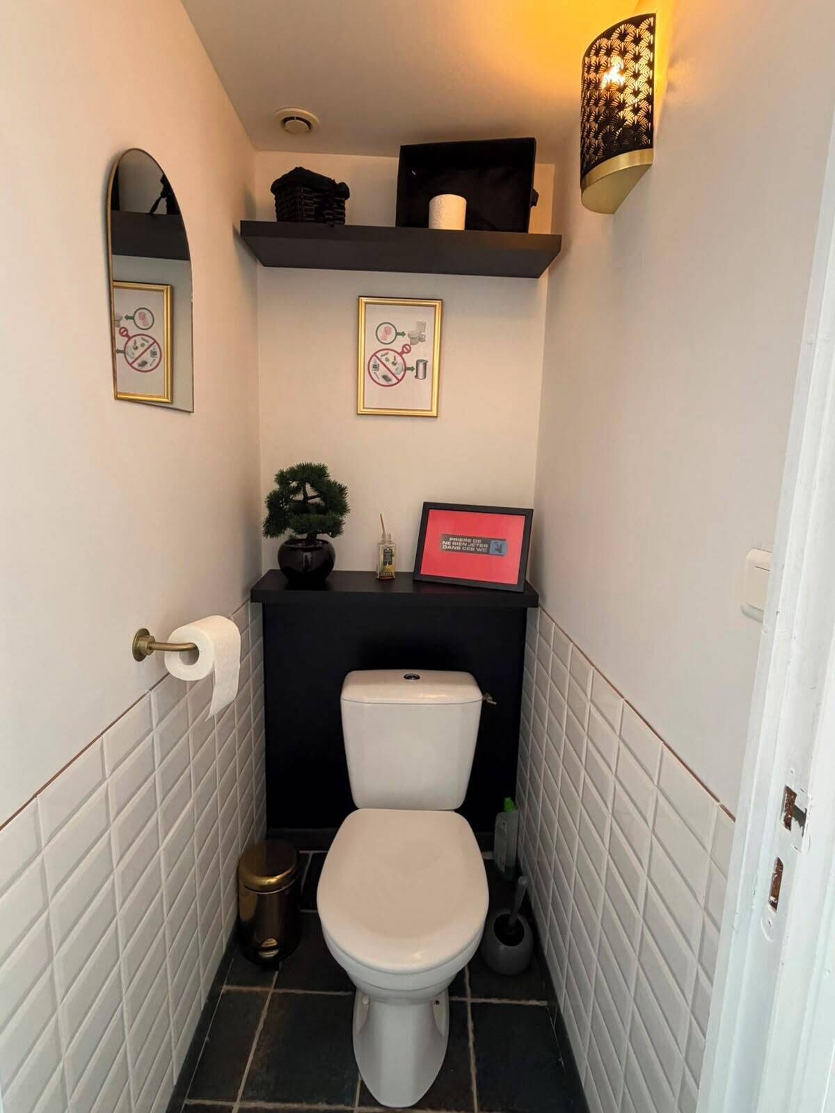 A compact bathroom with a toilet, a small shelf with toiletries, a framed picture, and a wall-mounted light fixture.