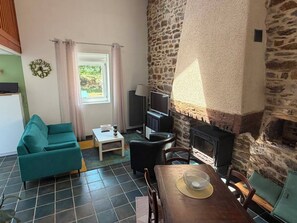 A cozy living room with a stone fireplace, a teal sofa, a small table, and a television. - Vacation Rental in the Brocéliande Forest (Néant-sur-Yvel)