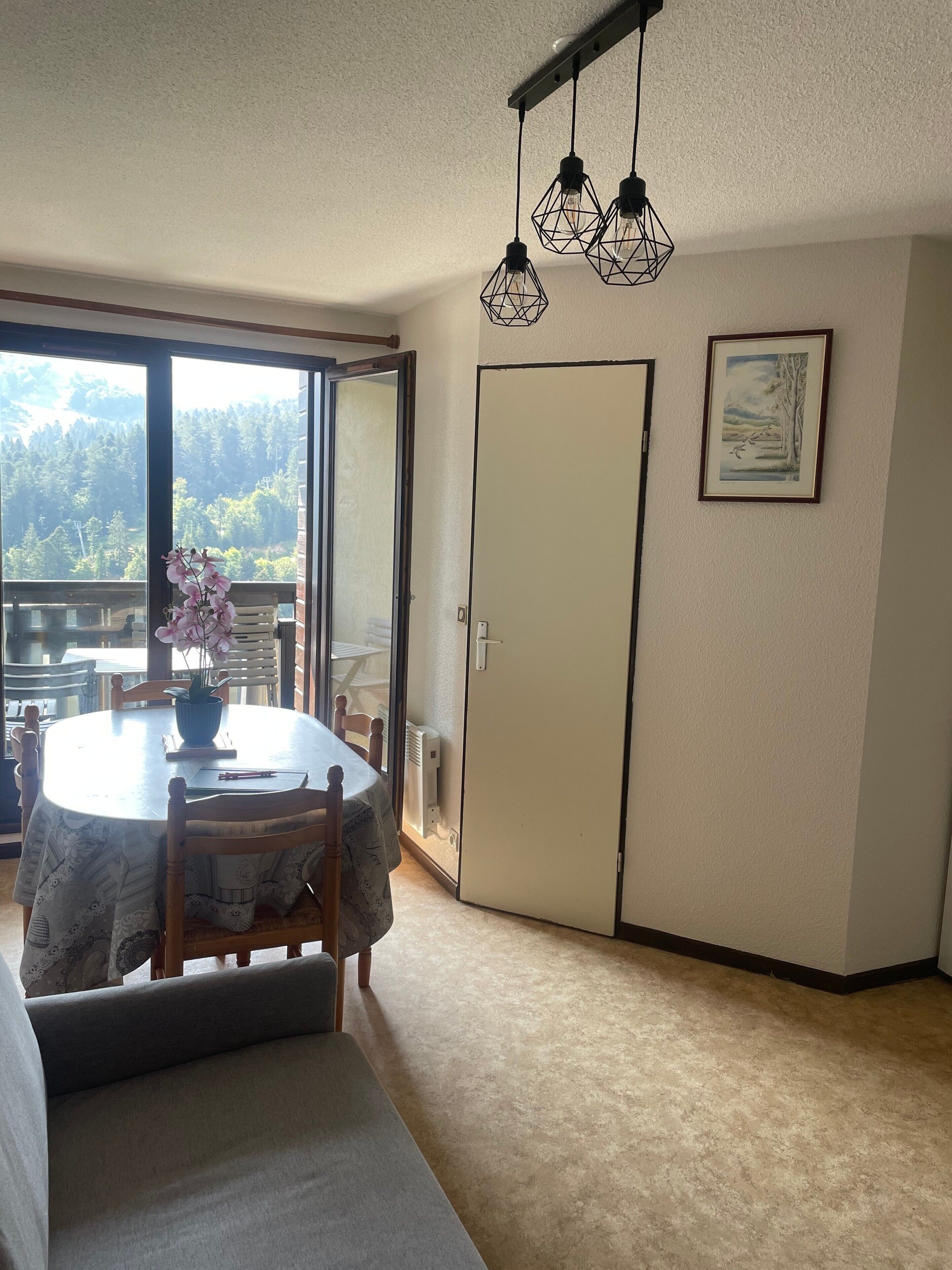 Un coin repas comprenant une table, des chaises, un vase de fleurs et une vue sur une forêt, visible à travers une porte-fenêtre coulissante.