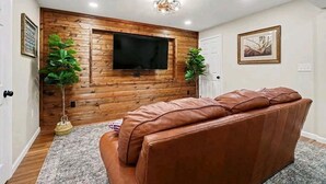 A living room with a brown leather sofa, wooden panel wall, mounted TV, and two potted plants. - Family Retreat, Hunting Lodge, or adventure base camp - The choice is yours (Blain)