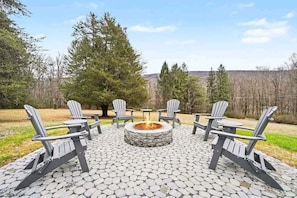 A fire pit surrounded by Adirondack chairs on a stone patio with a view of trees and hills.