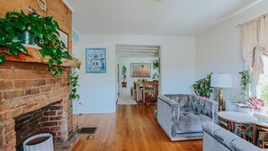 A living room with a brick fireplace, a grey sofa, wooden flooring, and a dining area in the background. - The Mariposa (Staunton)