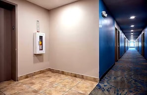 A hotel hallway with a fire alarm on the wall, a carpeted floor, and doors on both sides.